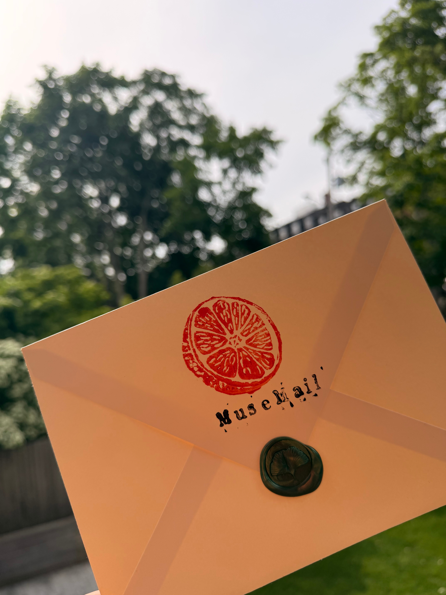 Cream Muse Mail envelope tied with twine and sealed with a green ginkgo wax stamp, with a hand-stamped orange slice and Muse Mail logo